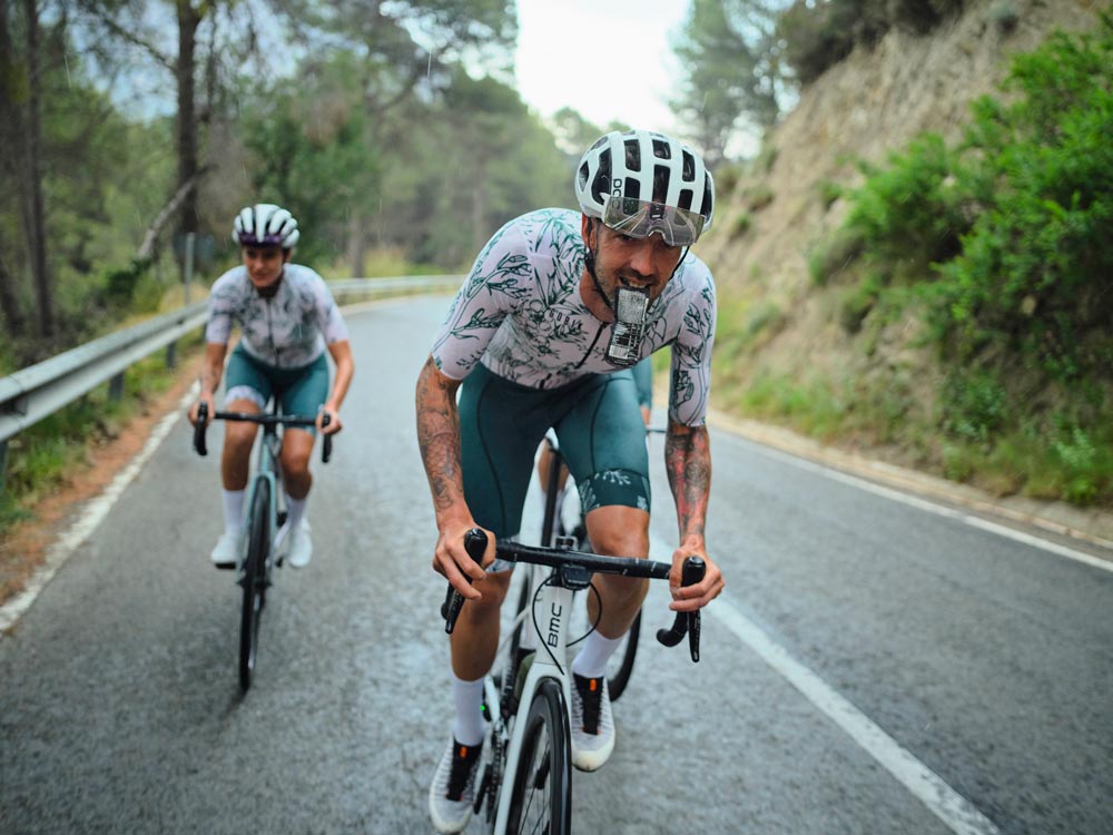 Un ragazzo e una ragazza dello stesso gruppo pedalano insieme su un passo di montagna, il ragazzo tiene in bocca un gel. Indossano Gobik maglia nelle tonalità chiare del verde e del bianco, con motivi floreali, e pantaloncini verdi anch'essi di Gobik pantaloncini.