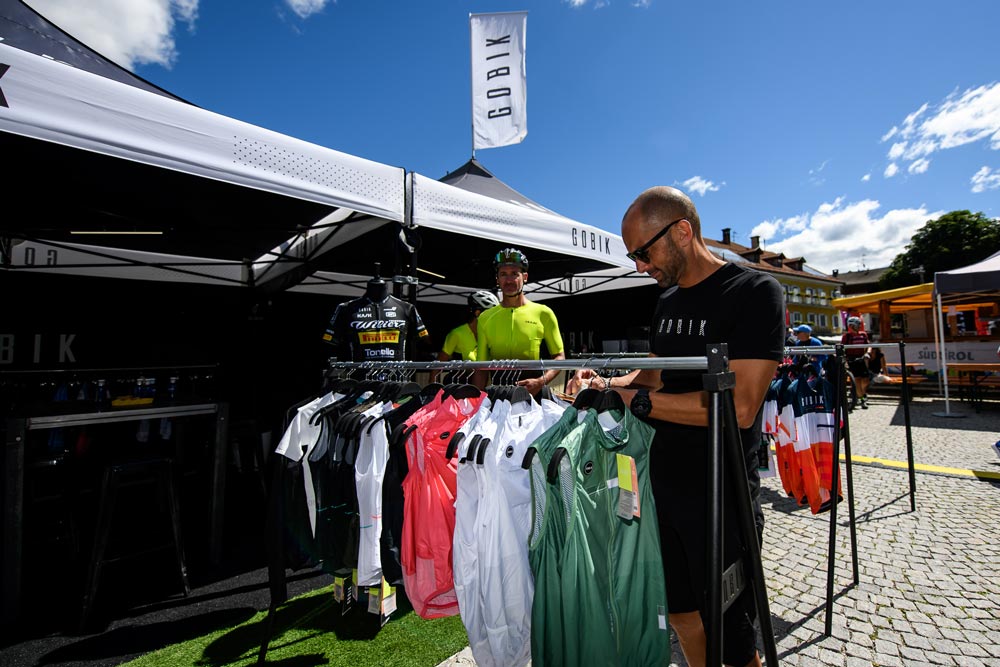 Miembro del staff de Gobik colocando las prendas en el expositor metálico dispuesto en la feria del corredor de un evento. Las prendas expuestas son chalecos de la marca en diferentes colores. Al fondo de la imagen, aficionados curiosos se acercan a la zona.