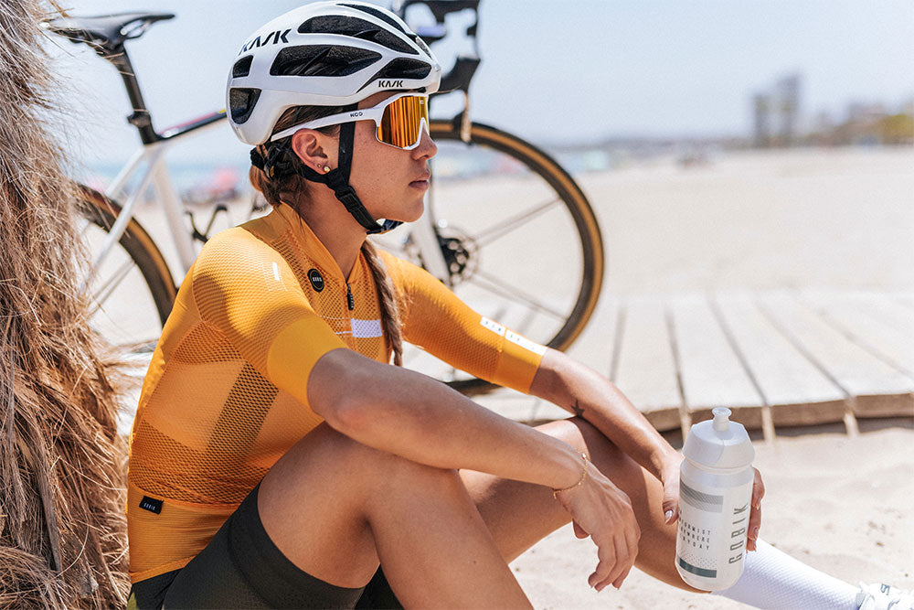 Cyclist girl drinks water from jerry can sitting on a beach in bright summer sunshine