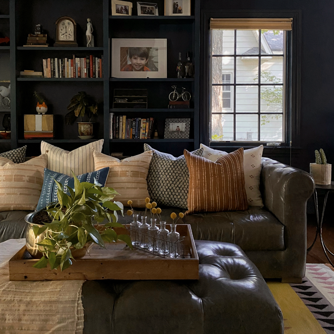 Dark living room with a leather couch filled with colorful throw pillows in front of a book shelf.