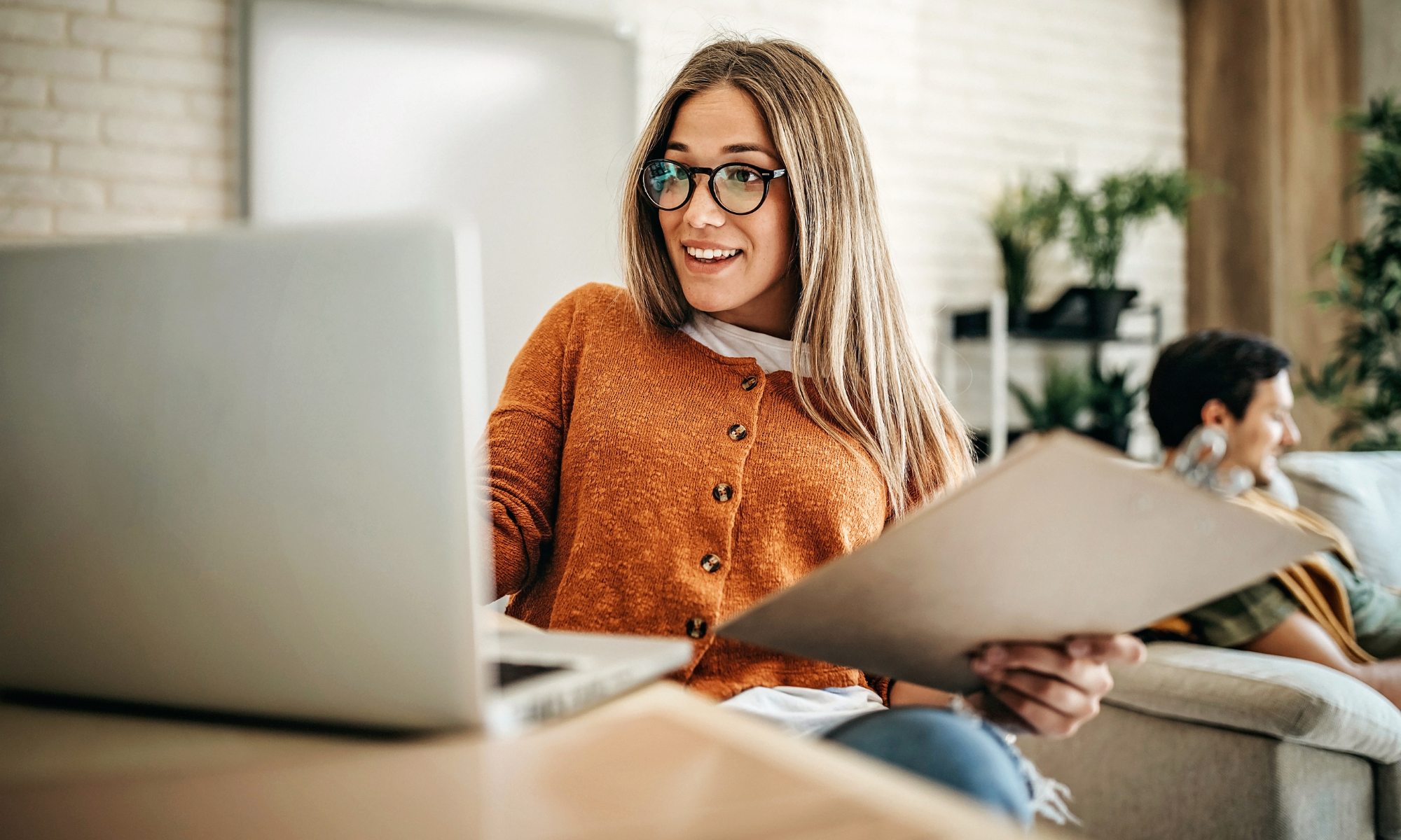 Young smiling professional holding manila folder in front of laptop sitting on living room table.