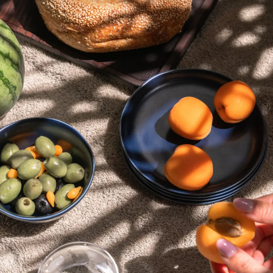 Apricots, olives, and bread arranged on a textured cloth in dappled sunlight.
