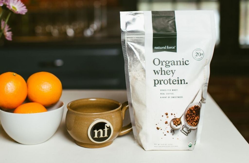 bag of natural force organic whey protein next to a handcrafted natural force mug beside a bowl of oranges