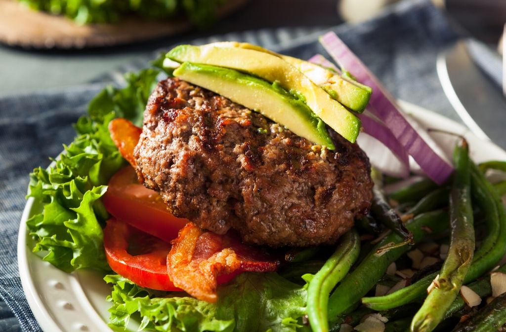 a white plate with lettuce tomato and green beans with a burger patty topped with avocado slices on top