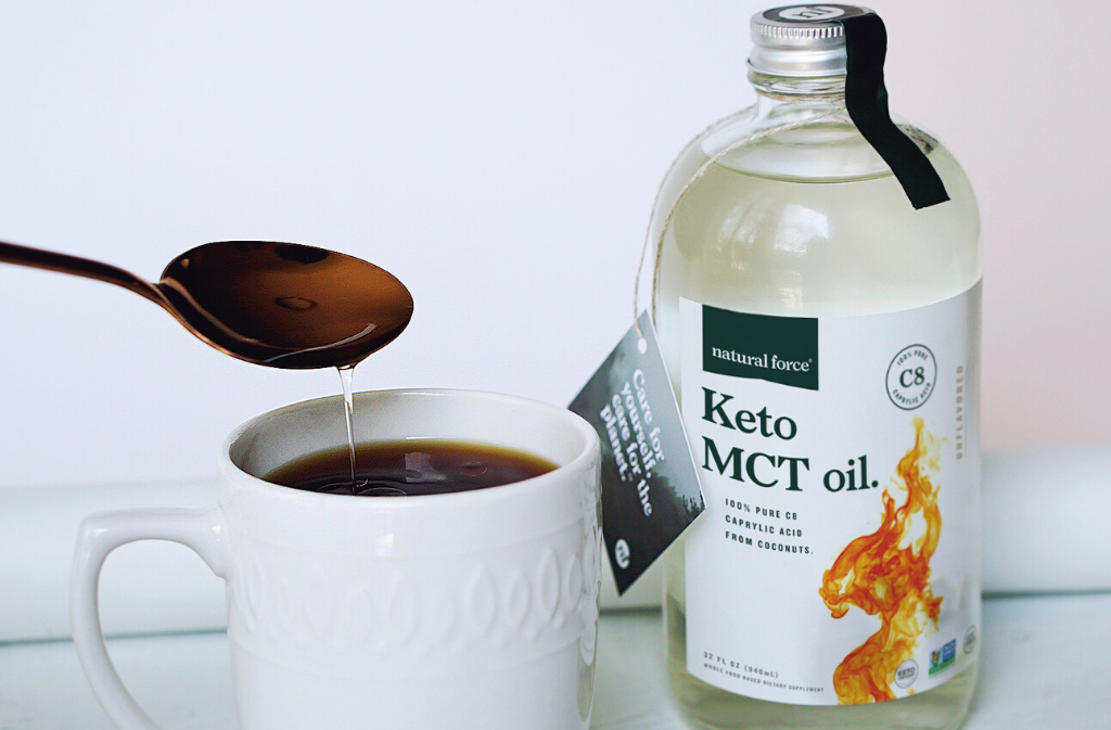 a bottle of natural force keto mct oil beside a white mug of coffee with keto mct oil being poured into it