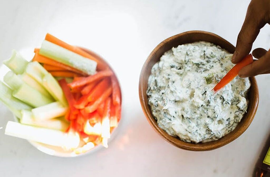 hand dipping a carrot stick into a bowl of keto spinach artichoke dip