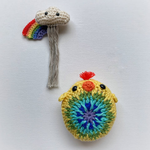 Crochet rainbow and rain cloud together with a rainbow coloured crochet chick against a white background