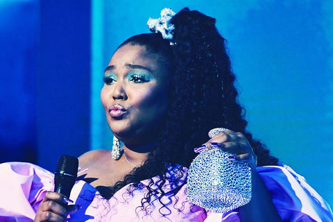 Lizzo performing on stage in an 80's style purple dress with a jewelled silver scrunchie and a blue background