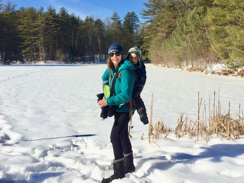A woman hiking in the snow wearing Columbia snowboots with a toddler on her back in an Onya Outback carrier