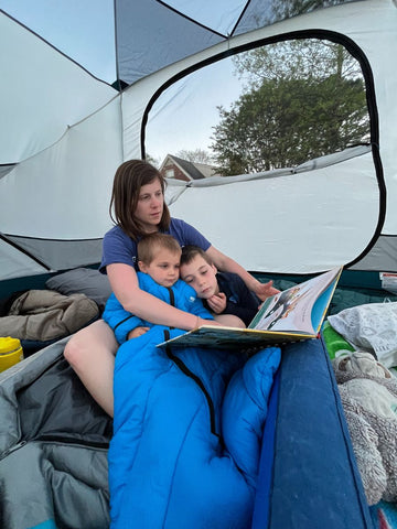 A woman sitting in a tent with two young boys wearing a Morrison Outdoors sleeping bag reading a book in a backyard