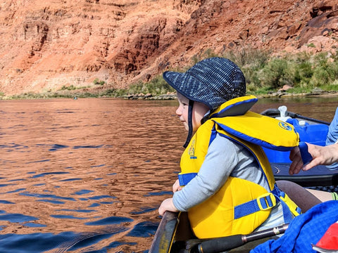 A baby in a canoe wearing a yellow life jacket, merino wool, and a sun hat floating along a river