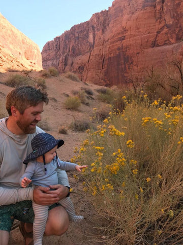 A man holding a baby who is touching yellow flowers in a scenic canyon area