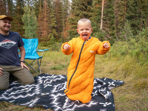 A man smiling as a young child makes a silly face while wearing a Morrison Outdoors Big Mo sleeping bag outdoors