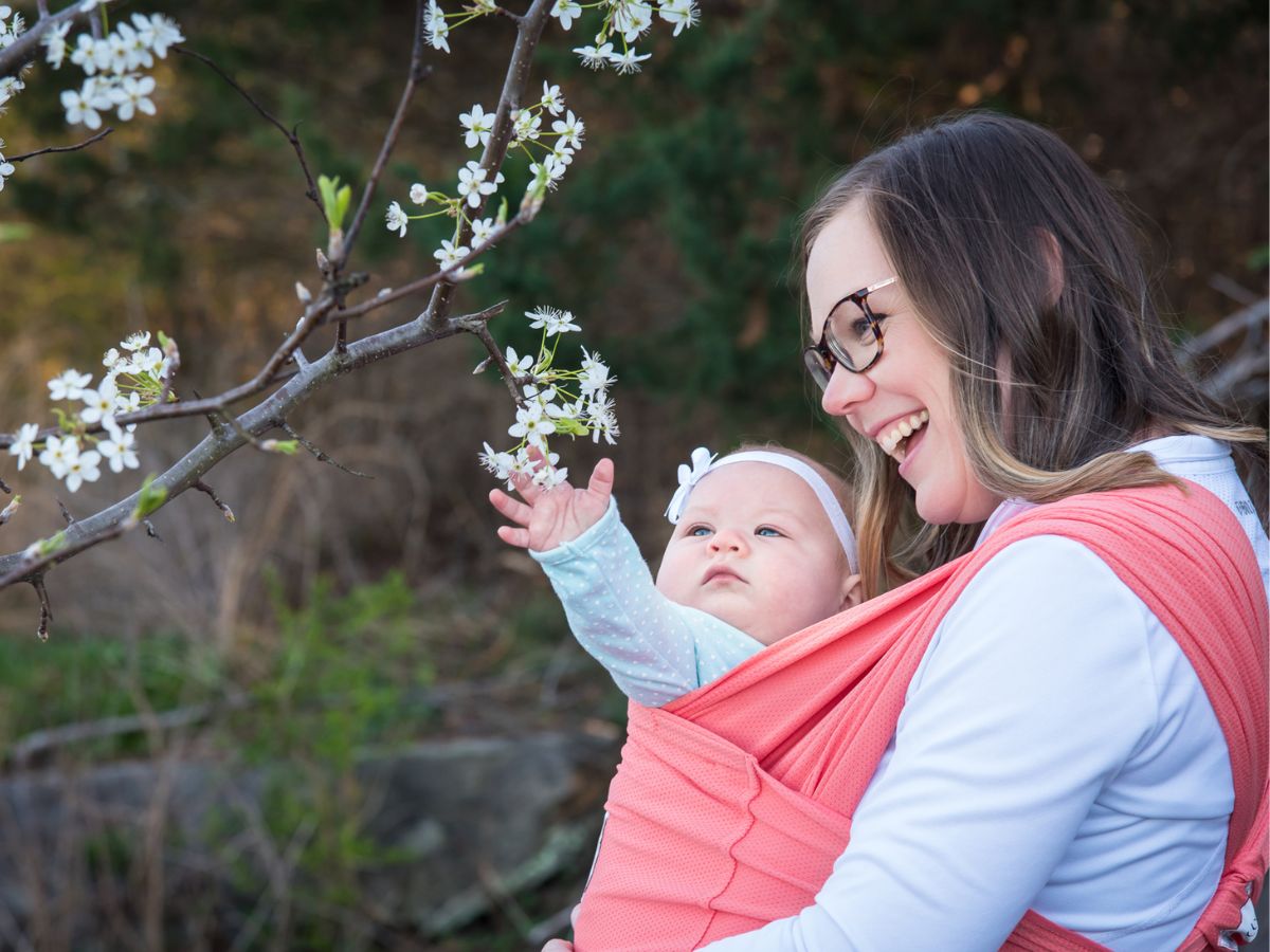 A woman carrying a baby in a pink wrap carrier in an outdoor setting while the baby reaches for flowers on a tree