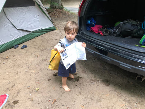A young boy carrying a map and wearing a backpack in front of a tent next to an SUV