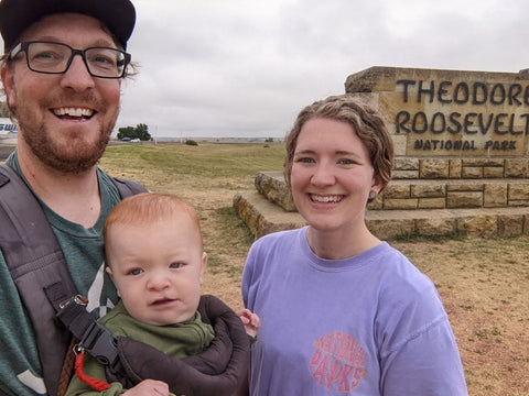 A woman and a man wearing a baby in a front-facing carrier standing in front of a sign for Theodore Roosevelt National Park