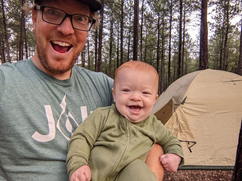 A man holding a baby while both smile in front of a tent