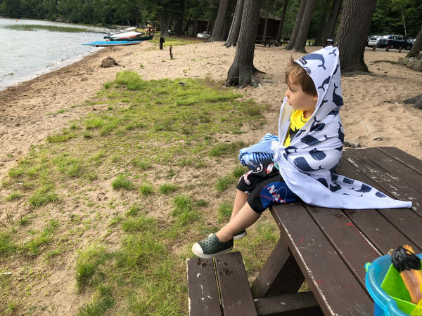 A young boy sitting on a picnic table wrapped in a towel while watching the water at a campground beach