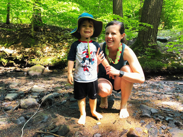 A woman standing in a shady creek with a young boy who is wearing a sun hat and sun shirt