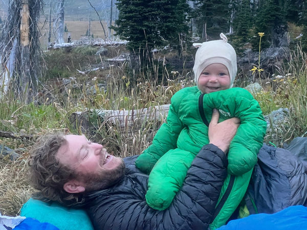 A man holding a smiling baby wearing a Morrison Outdoors Little Mo Sleeping Bag while camping