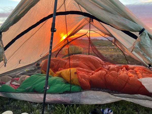 An adult and a baby in a Morrison Outdoors Little Mo Sleeping Bag sleeping in a tent with a view of the sunrise in the background
