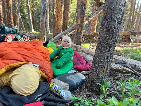 A baby wearing a Morrison Outdoors Little Mo sleeping bag sitting next to a sleeping adult in a forested area