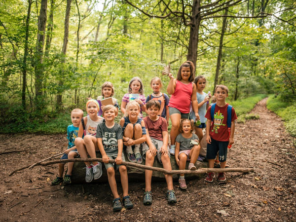 A group of children smiling and sitting on a log on a forested trail