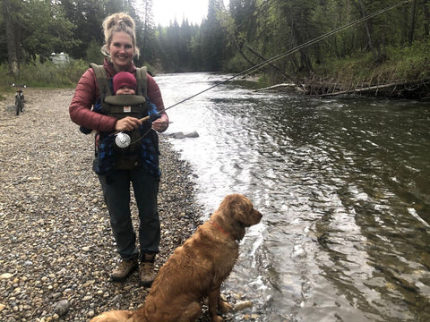 A woman fly fishing near a river with a baby in a baby carrier and a golden retriever at her feet