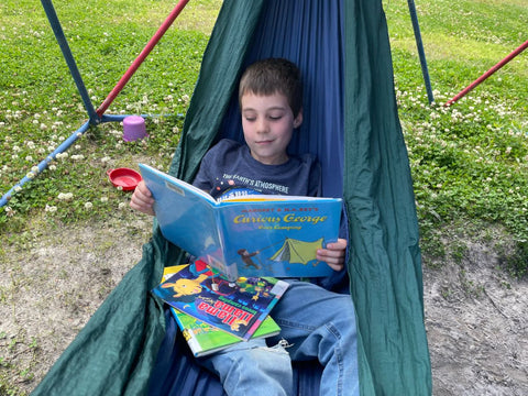 A young boy reading Llama Llama Loves Camping in a hammock in a backyard
