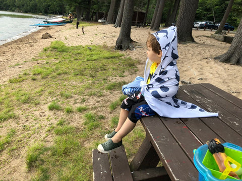 A young boy sitting on a picnic table wrapped in a towel looking out at the water on a lakeside beach