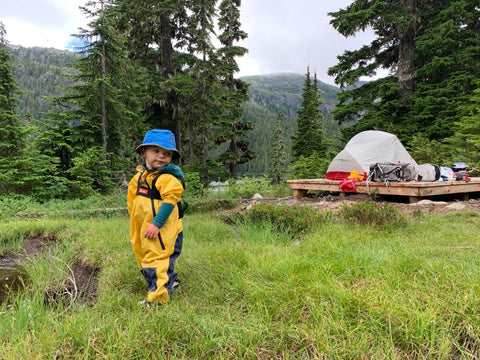 A toddler in a yellow rainsuit exploring a grassy area in front of a camping platform with a tent