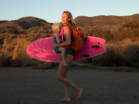 A woman carrying a bookie board and a child along the beach in a Trail Magik child carrier attached to a hiking backpack