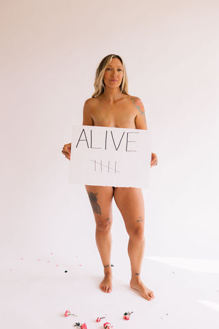 Breast Cancer Survivor Holly is Standing & Posing with a sign that reads "ALIVE".