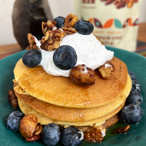 A stack of low carb almond flour pancakes made with Cinnamon Vanilla 5 Nut Granola, surrounded by blueberries, more granola, and topped with whipped cream.