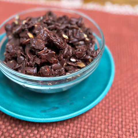 A bowl of sugar free granola bites made with Better Than Provisions 5 Nut Granola inside a clear bowl sitting on top of a teal blue plate. A rust colored tablecloth is beneath it.