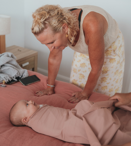 A mother peering over a baby who is swaddled. Baby happy and content. Swaddle is a rose colour.