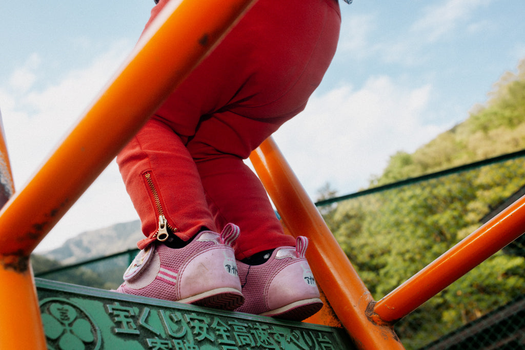 An image taken for NiMi Projects UK of a child's legs in red trousers and pink shoes as she sits on a climbing frame in Japan. 
