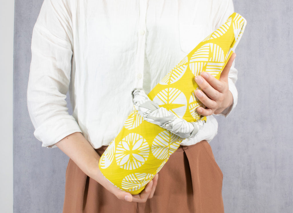 A woman holding a large bottle wrapped in a yellow and grey reversible patterned furoshiki wrapping cloth.