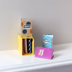 Dotted, pink and blue origami minature notebooks made with dotted paper, displayed around and inside a miniature yellow origami bookshelf in a photo for NiMi Projects Origami Workshops.