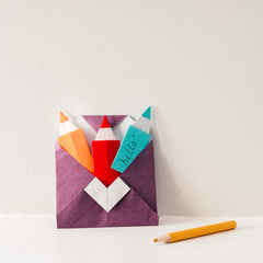 A purple origami envelope containing three origami pencil shaped notelets in orange, red and blue, displayed with a pencil, in a photo taken by NiMi Projects for its origami workshops.