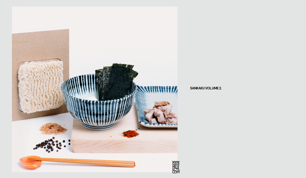 A photo of the hardback version of Sankaku Vol. 2: Ramen book, featuring a vacuum packed block of ramen noodles as part of its cover design, on display with a blue striped noodle bowl and a matching plate.