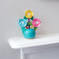 Three origami roses, in blue, yellow and pink with green origami leaves and colored popsicle stick stems, displayed in a turquoise Okada Japanese ceramic round vase on a white shelf. Photo taken by NiMi Projects for its origami workshops. 