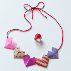 A necklace of origami hearts in pink, multicolored dotted, metalic pink, red striped and purple on a read thread, plus a matching red heart origami ring, made for a NiMi Projects origami workshop.