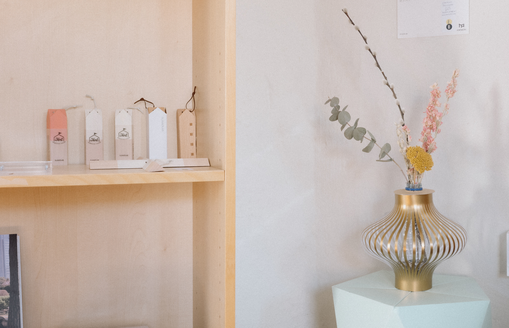 A shelf displaying NiMi Projects Sunao Lab House Trivets, next to a pale green Catachi origami polystool with a gold Fukunaga paper vase with flowers on top. Both on show at Chiddingstone Castle's gift shop in Kent, UK. 