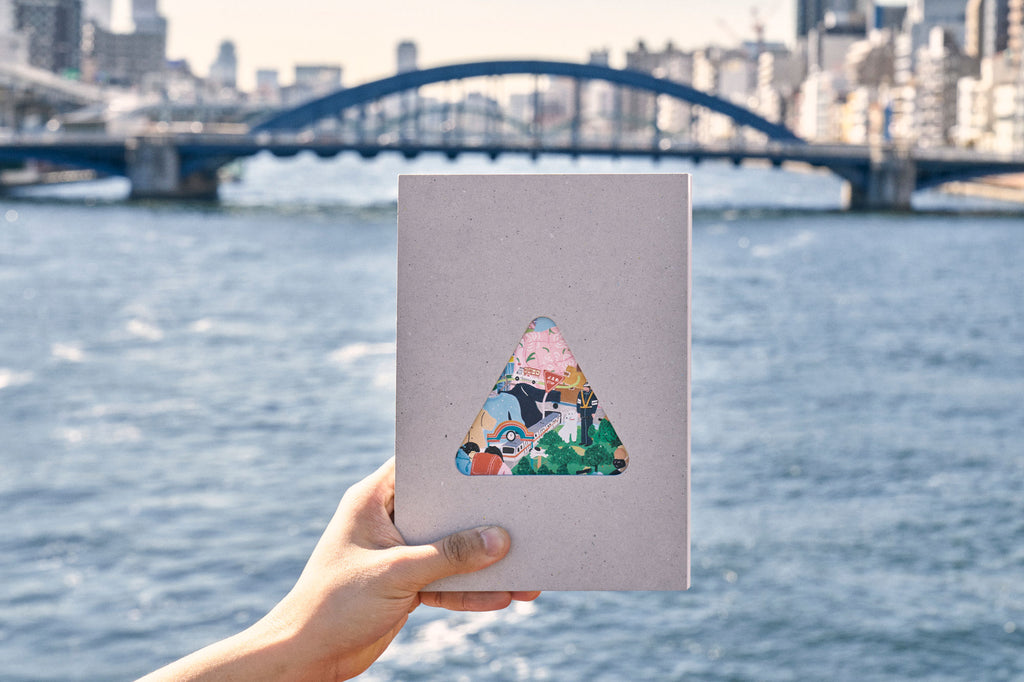 A copy of sankaku Vol. 1, a book featuring interviews with Tokyo craftspeople and creators, held up ahead of a bridge in Tokyo. The book's cover is grey with a central triangular cutout revealing a colourful illustration by Grace Lee.