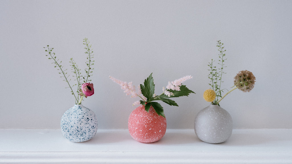 Three round Moheim Colour Drops bud vases, each with flowers and leaves, on a white shelf at NiMi Projects UK. From left to right, white with blue flecks, red with white flecks and grey with white flecks.