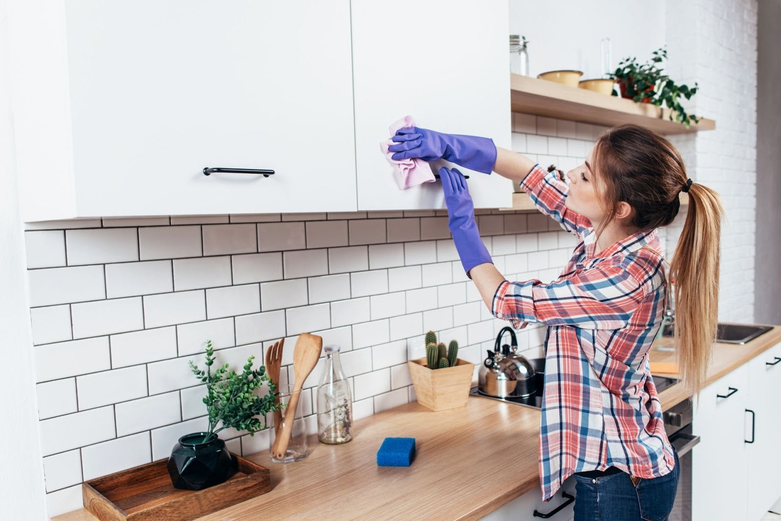 A woman in a plaid shirt and purple rubber gloves wiping kitchen cabinets with a microfibre cloth
