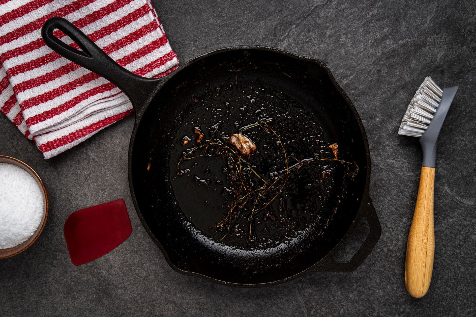 A dirty cast iron skillet, a brush, a dishcloth, and a bowl of salt on a kitchen counter