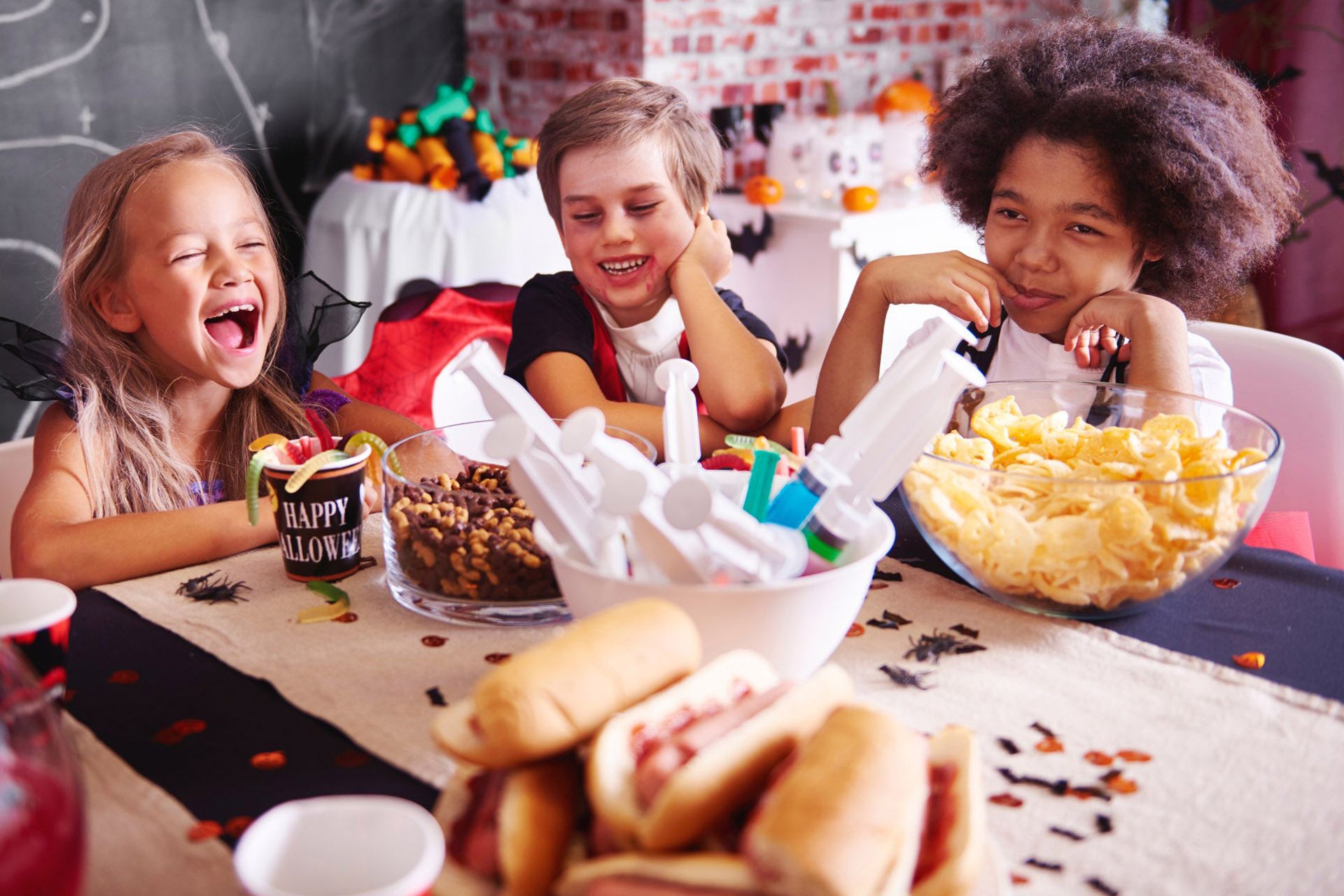 Three children laughing while enjoying Halloween candy at a festive snack table