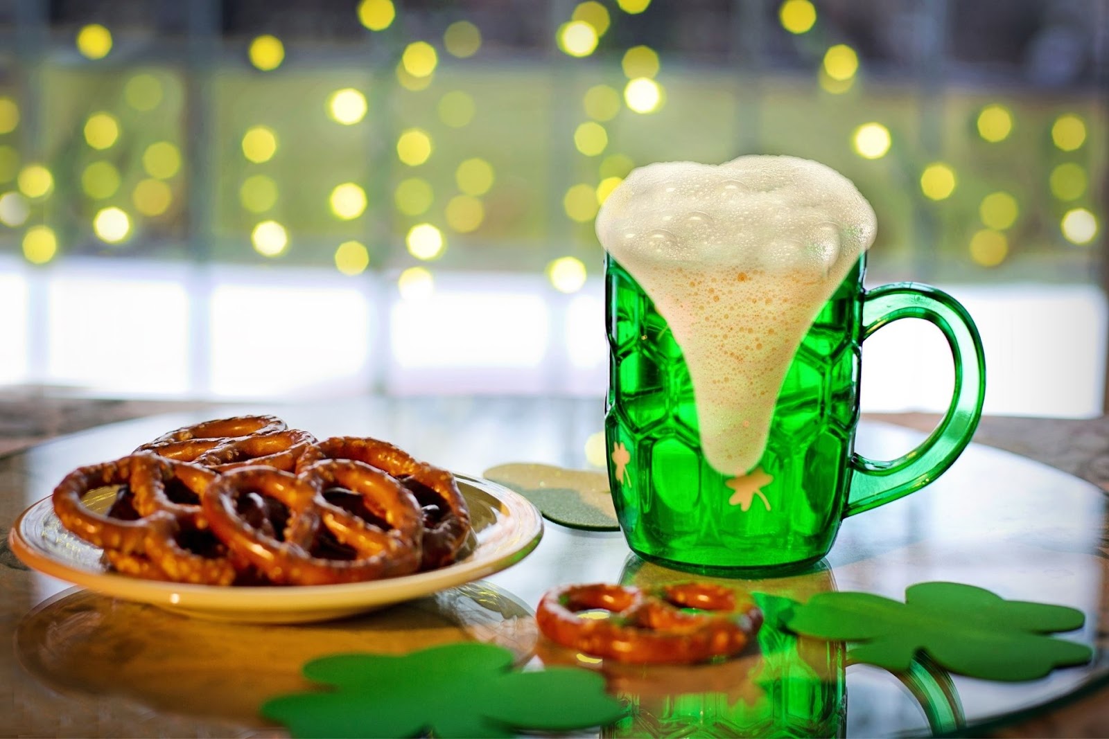 Pretzels, green beer, and shamrocks on a wooden table in front of gold fairy lights